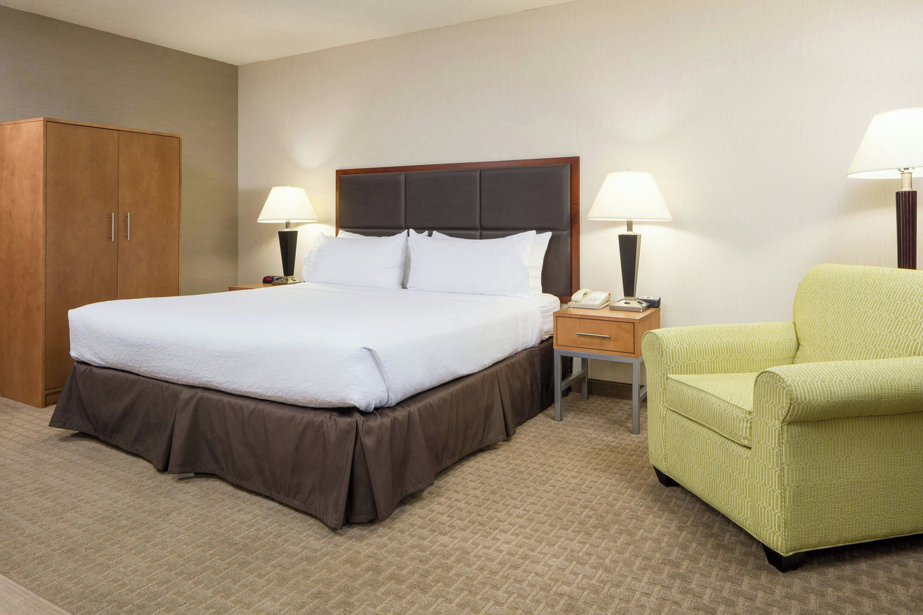 A hotel room with a bed, bedside lamps, a chair, a nightstand, and a wardrobe.
