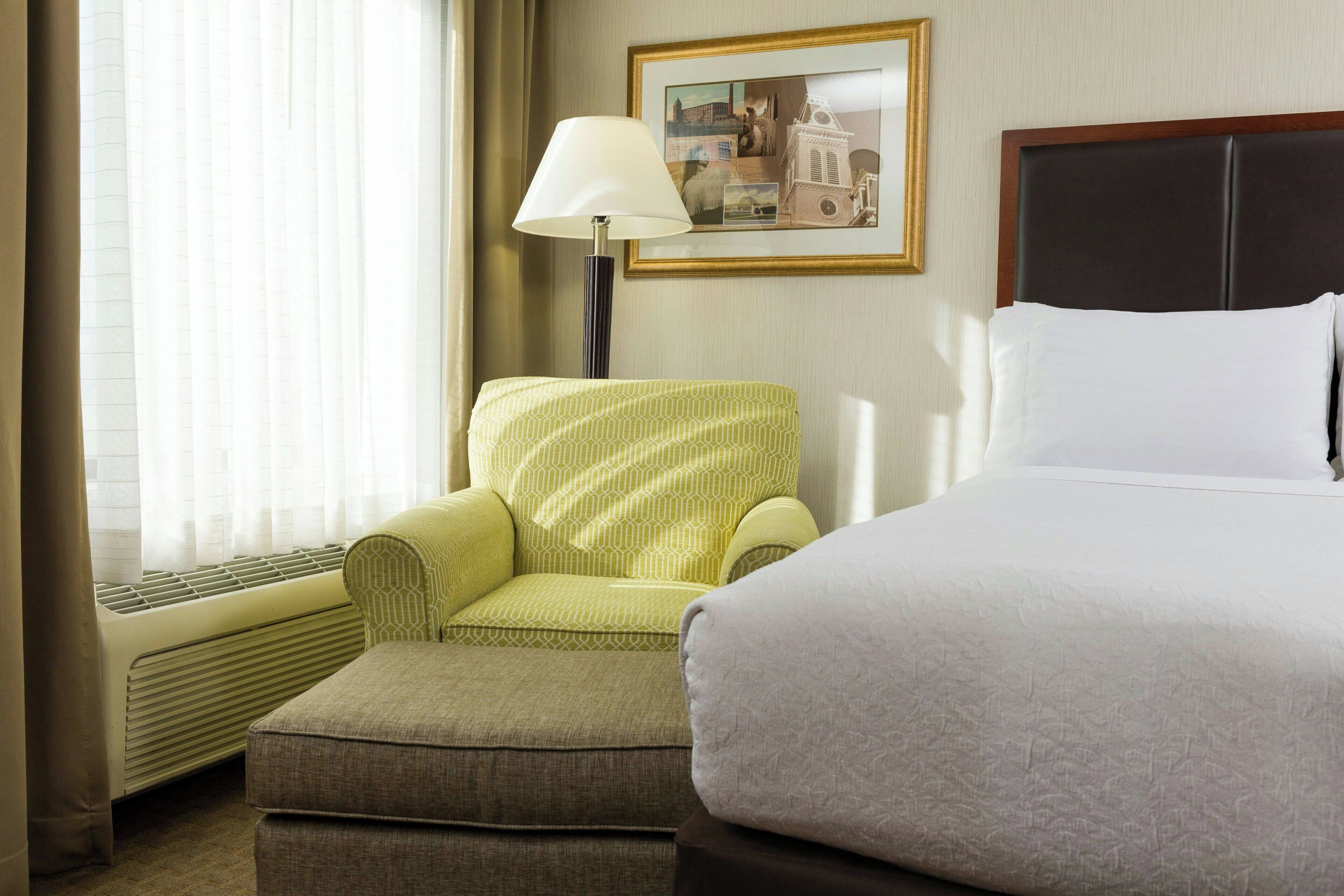 Una habitación de hotel con una cama, un sillón verde, un pequeño reposapiés, una lámpara de pie y un cuadro en la pared.