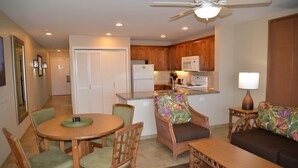 A compact kitchen with wooden cabinets, a microwave, and a refrigerator. There is a dining area with a round table and chairs, and a sofa with a floral cushion. - Sea Village (Kailua-Kona)