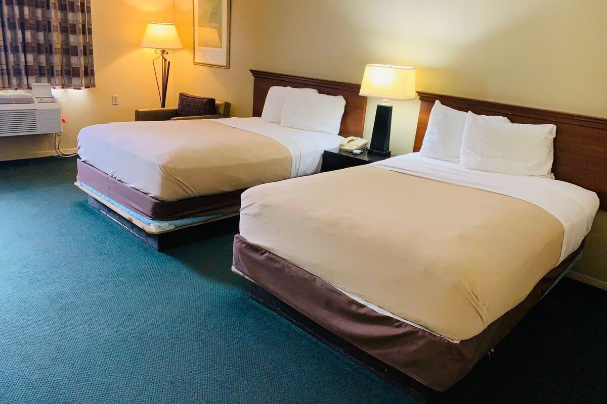 Habitación de hotel con dos camas, cada una con ropa de cama blanca y cabeceros de madera. Hay un escritorio con una lámpara, un teléfono y una computadora portátil. La habitación tiene una alfombra azul y una ventana con cortinas.
