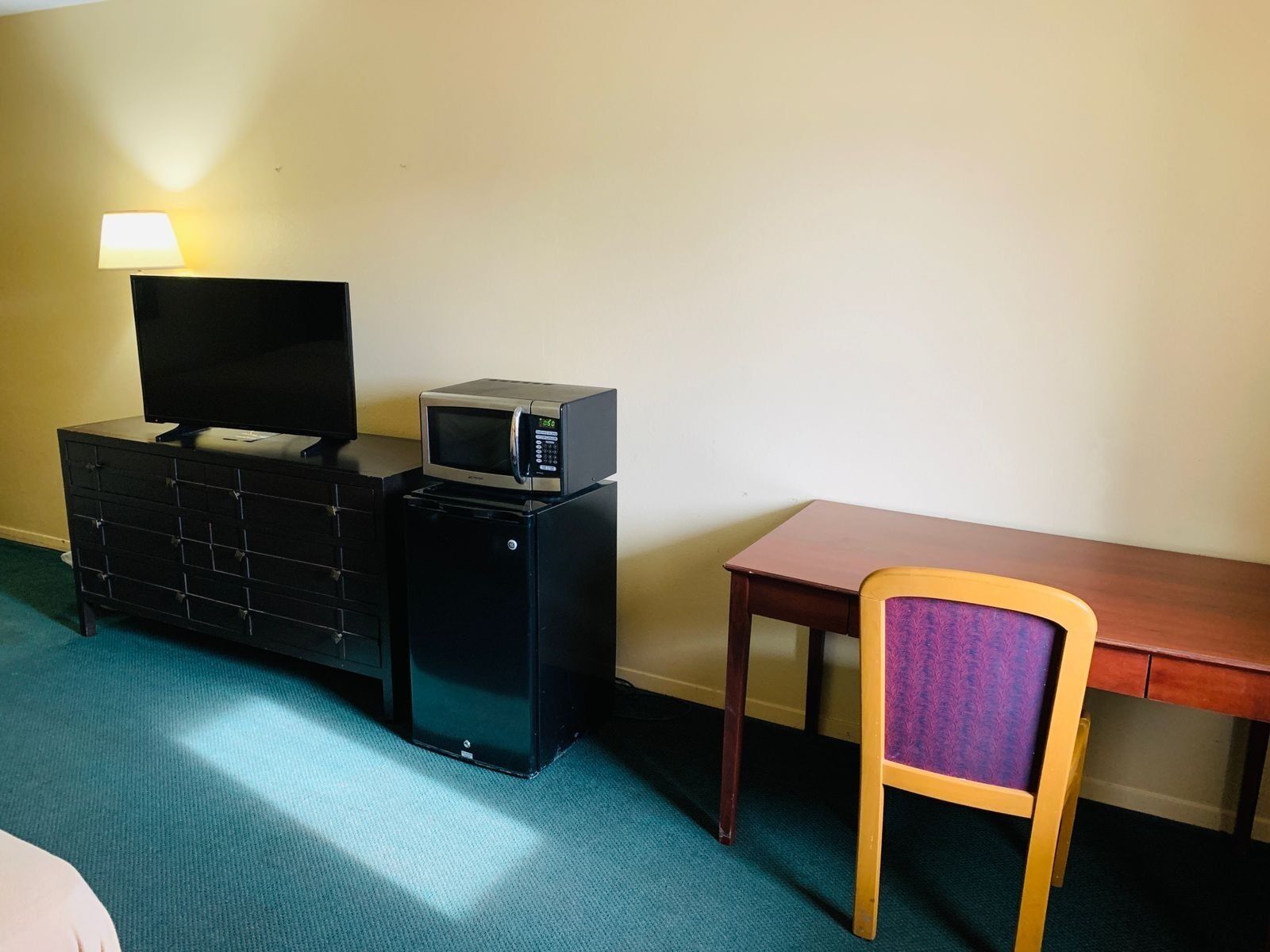 A hotel room with a TV on a dresser, a microwave on a small cabinet, and a wooden chair with a purple cushion.