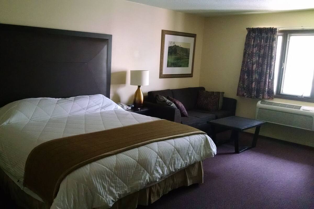 Habitación de hotel con una cama grande, un sofá pequeño, una mesita de noche con lámpara y una ventana con cortinas.