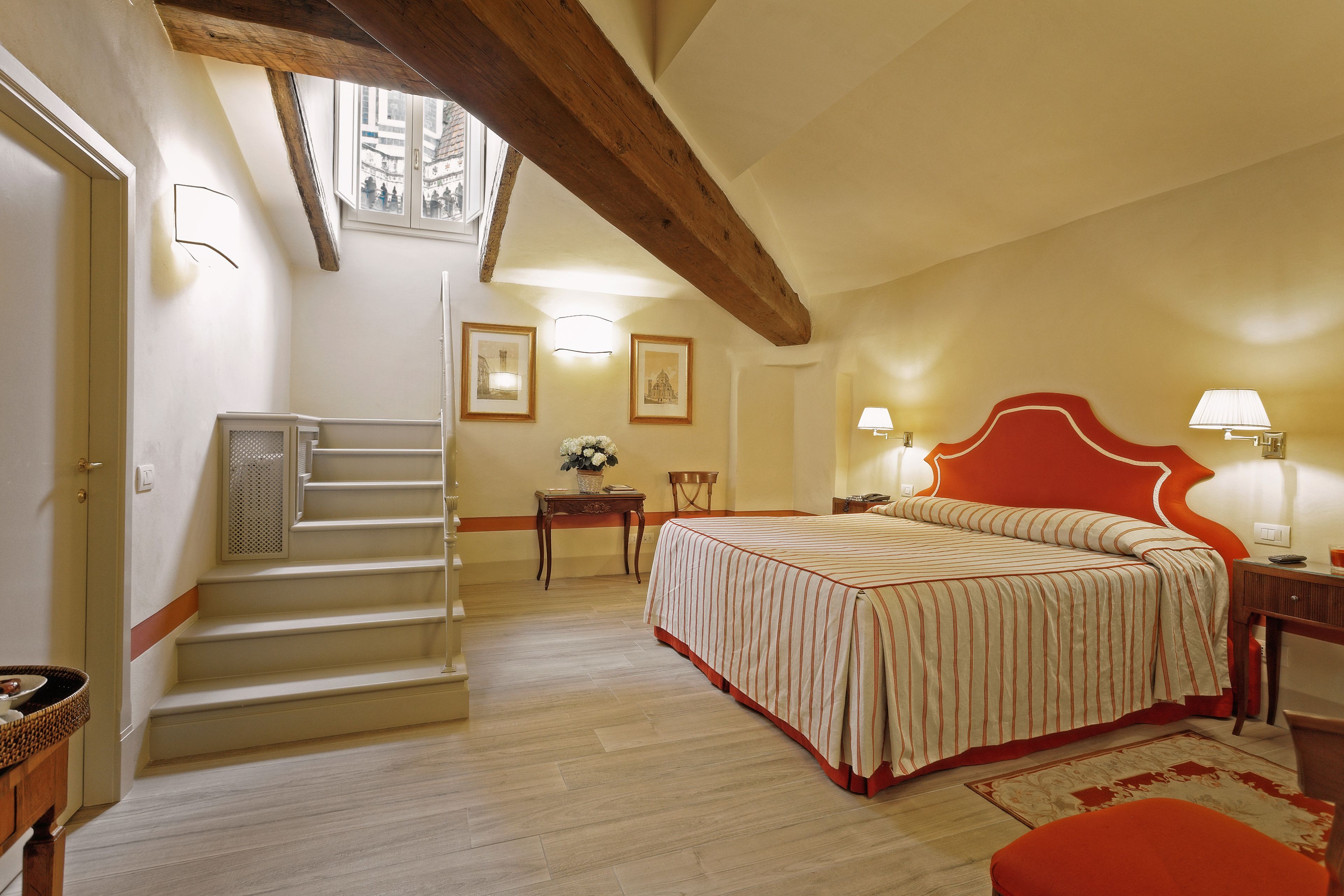A bedroom with a bed, a nightstand, a lamp, a chair, a small table with a vase, and a staircase.