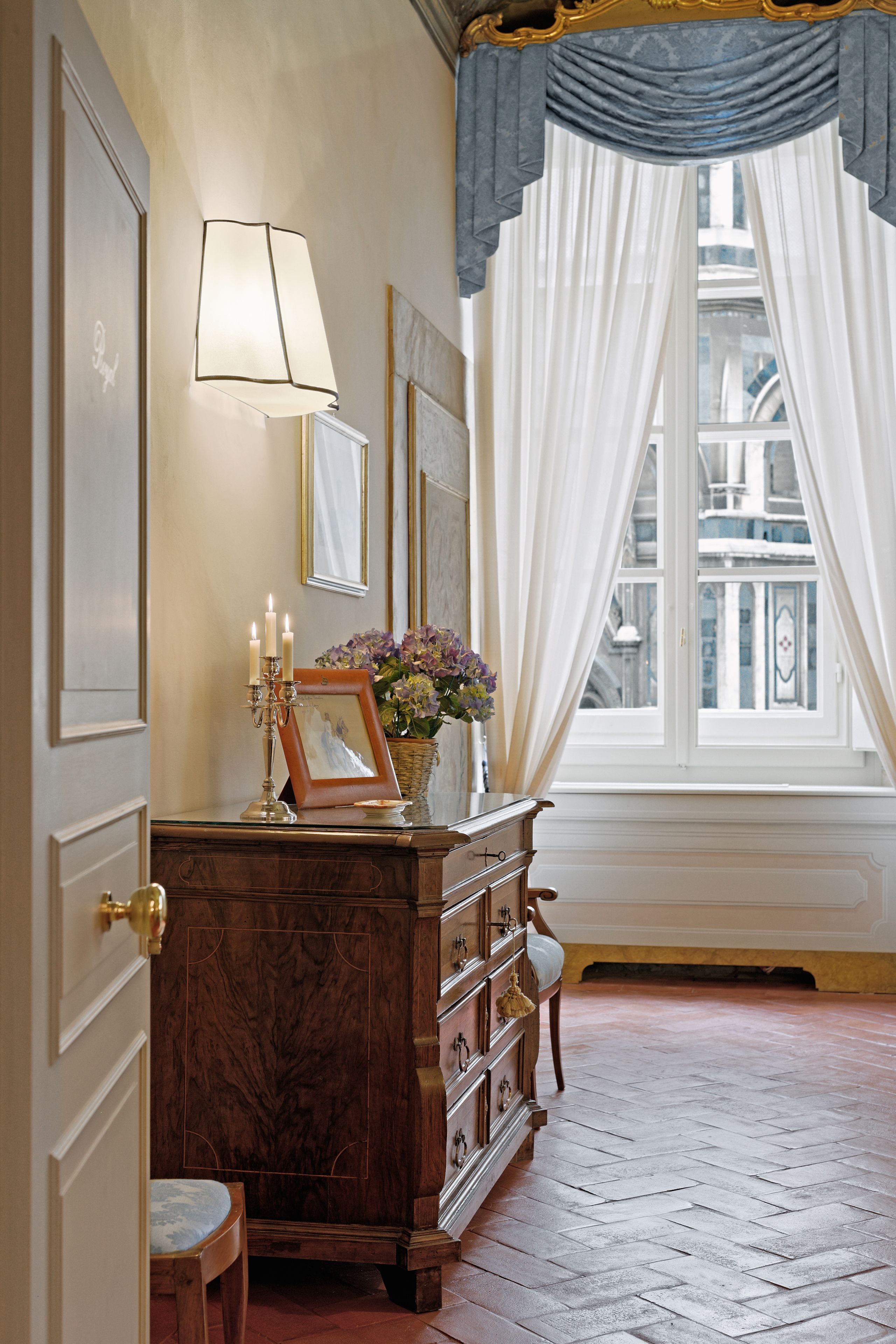 Une pièce comprenant une commode en bois, une lampe, un tableau encadré et une fenêtre ornée de rideaux légers.