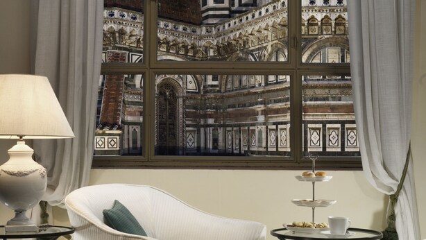 A room with a large window overlooking a historic building, a white chair, a small table with a lamp, and a tray with pastries. - Palazzo Niccolini al Duomo (Florence)
