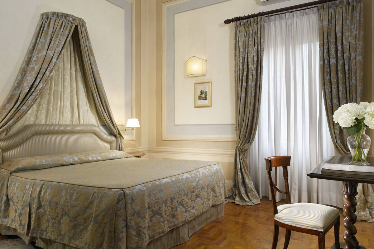 A bedroom with a four-poster bed, a chair, a table with a vase, and curtains.