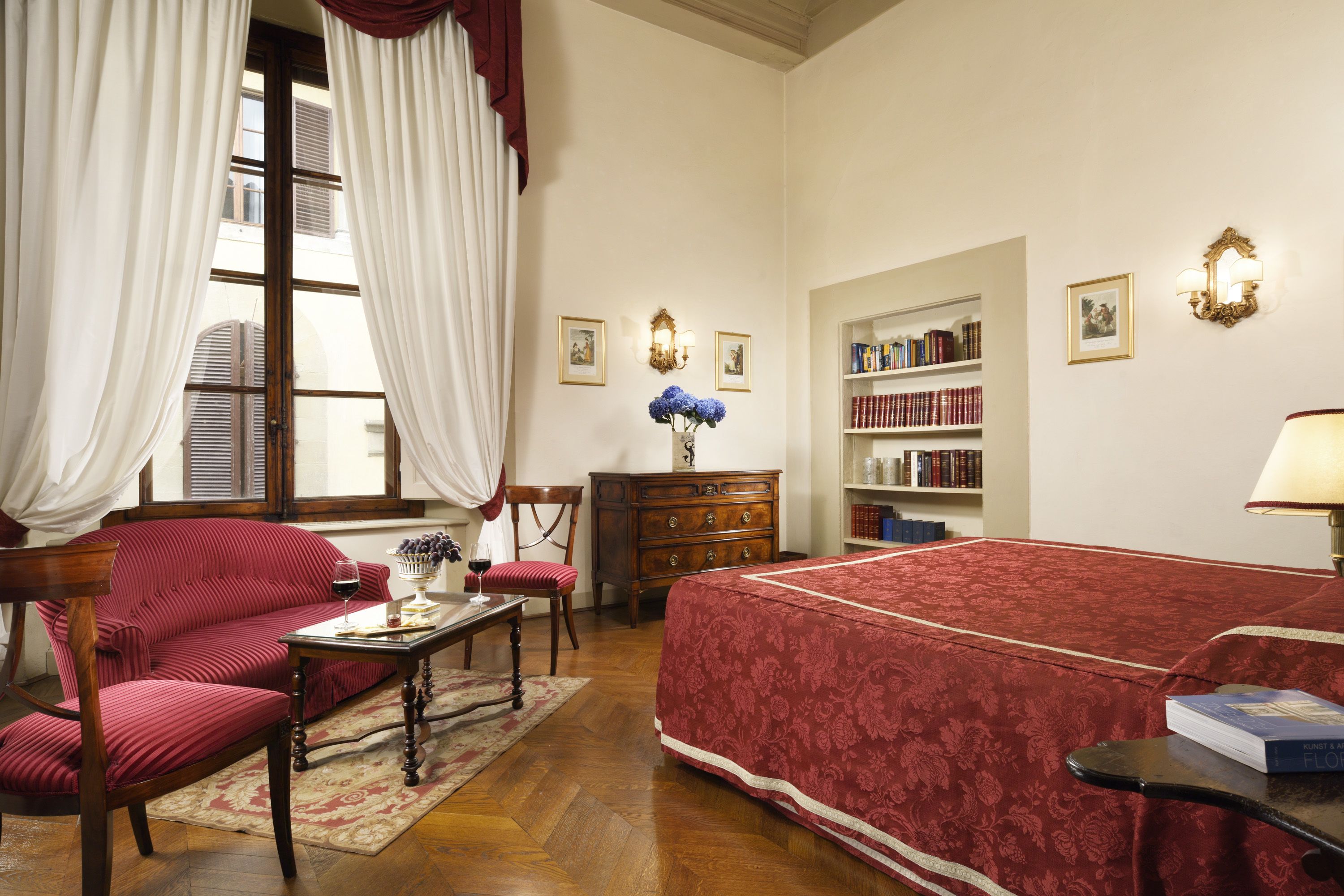 Une pièce avec un canapé rouge, une table basse en bois, une couvre-lit rouge, une bibliothèque et une fenêtre avec des rideaux.