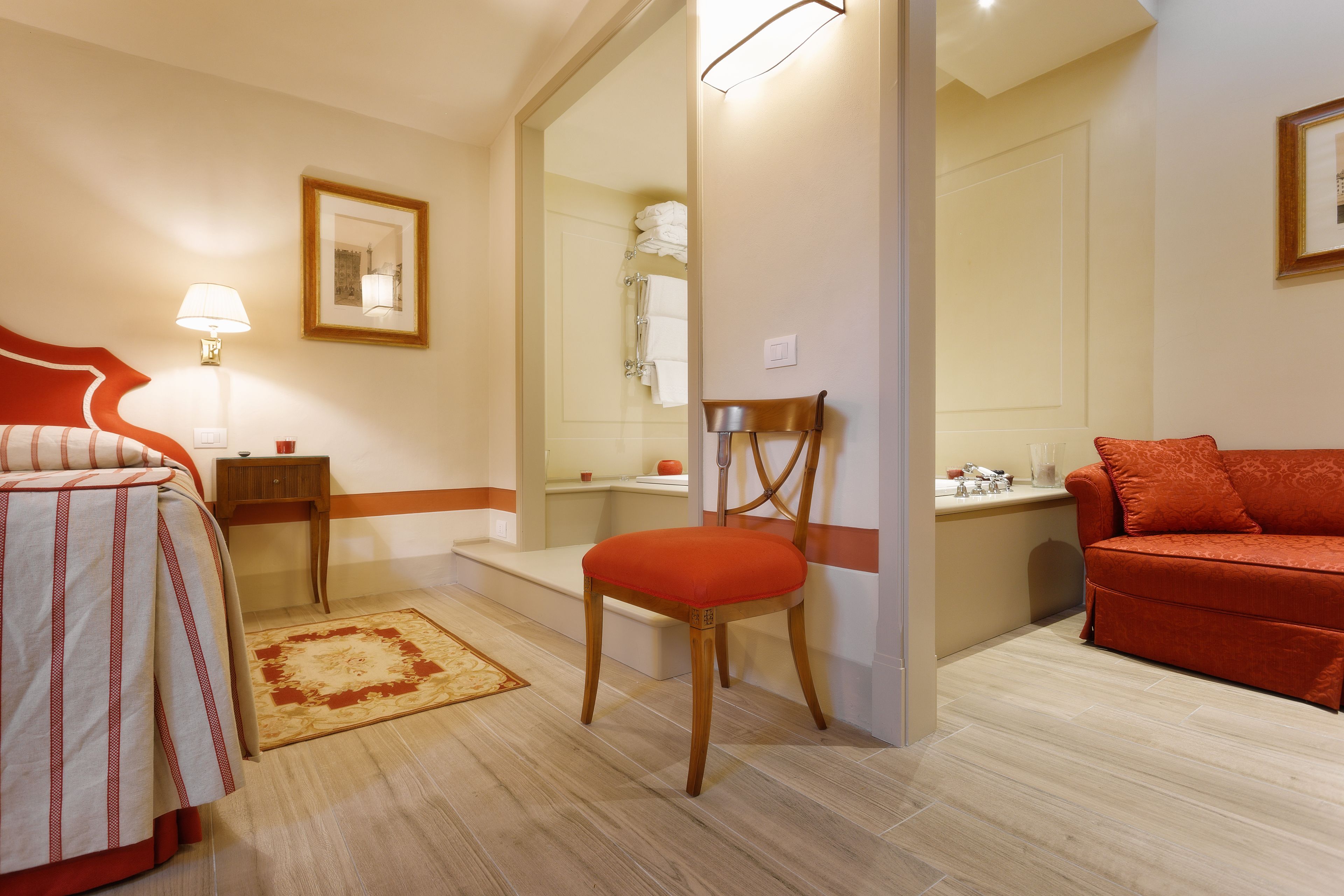 A hotel room with a bed, a chair, a sofa, a small table, a nightstand, a mirror, and a bathroom visible through a glass door.