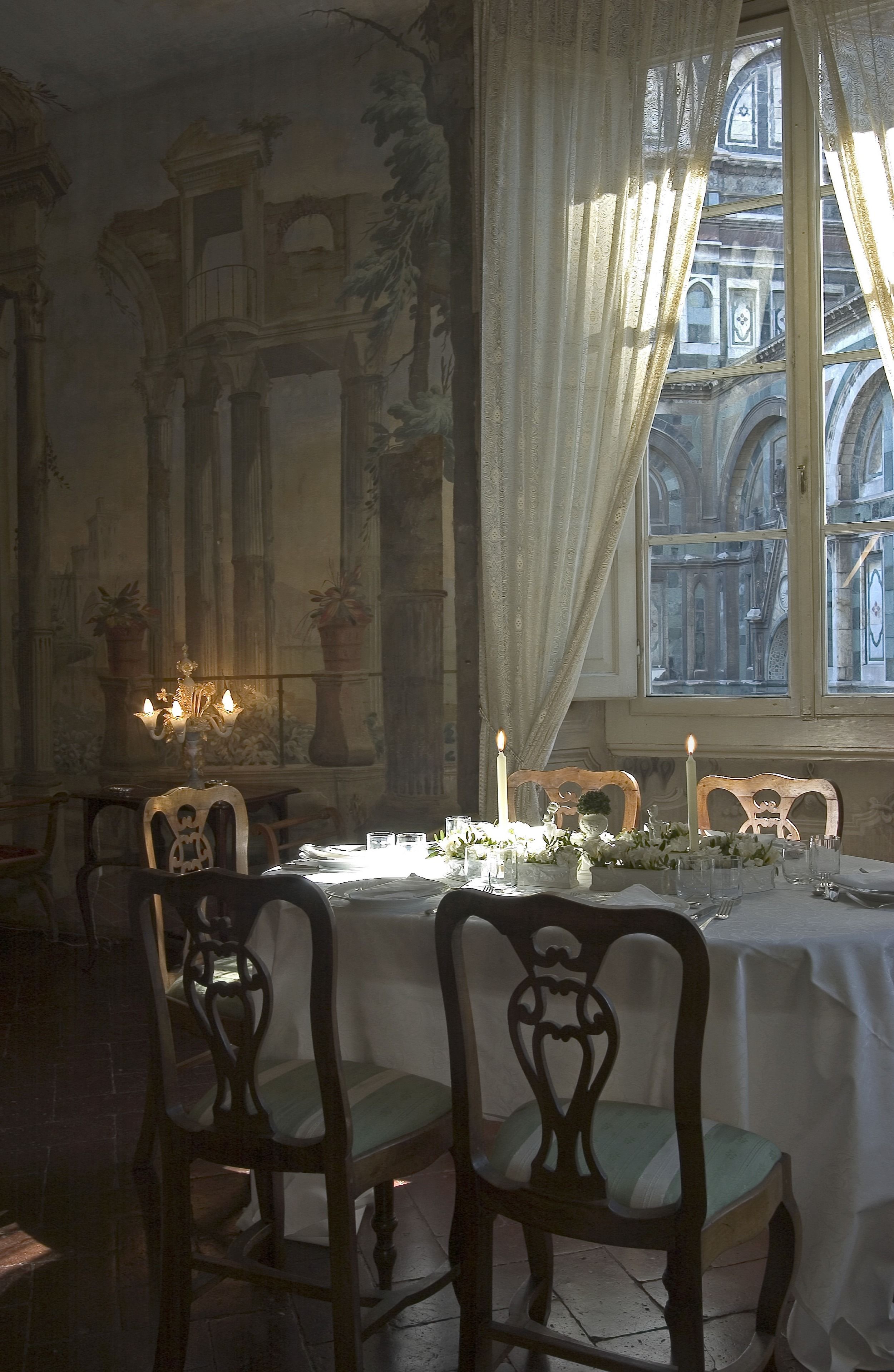 Une salle à manger avec une table dressée pour quatre personnes, des chaises décorées, un lustre et une vue sur un bâtiment historique par la fenêtre.
