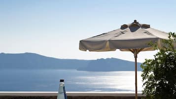 A table with a bottle, glasses, and a bowl of ice cubes, set under a large umbrella, overlooking a body of water with distant mountains.