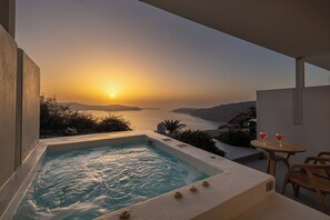 A hot tub with a view of the sunset over the sea, a table with a drink, and a chair.