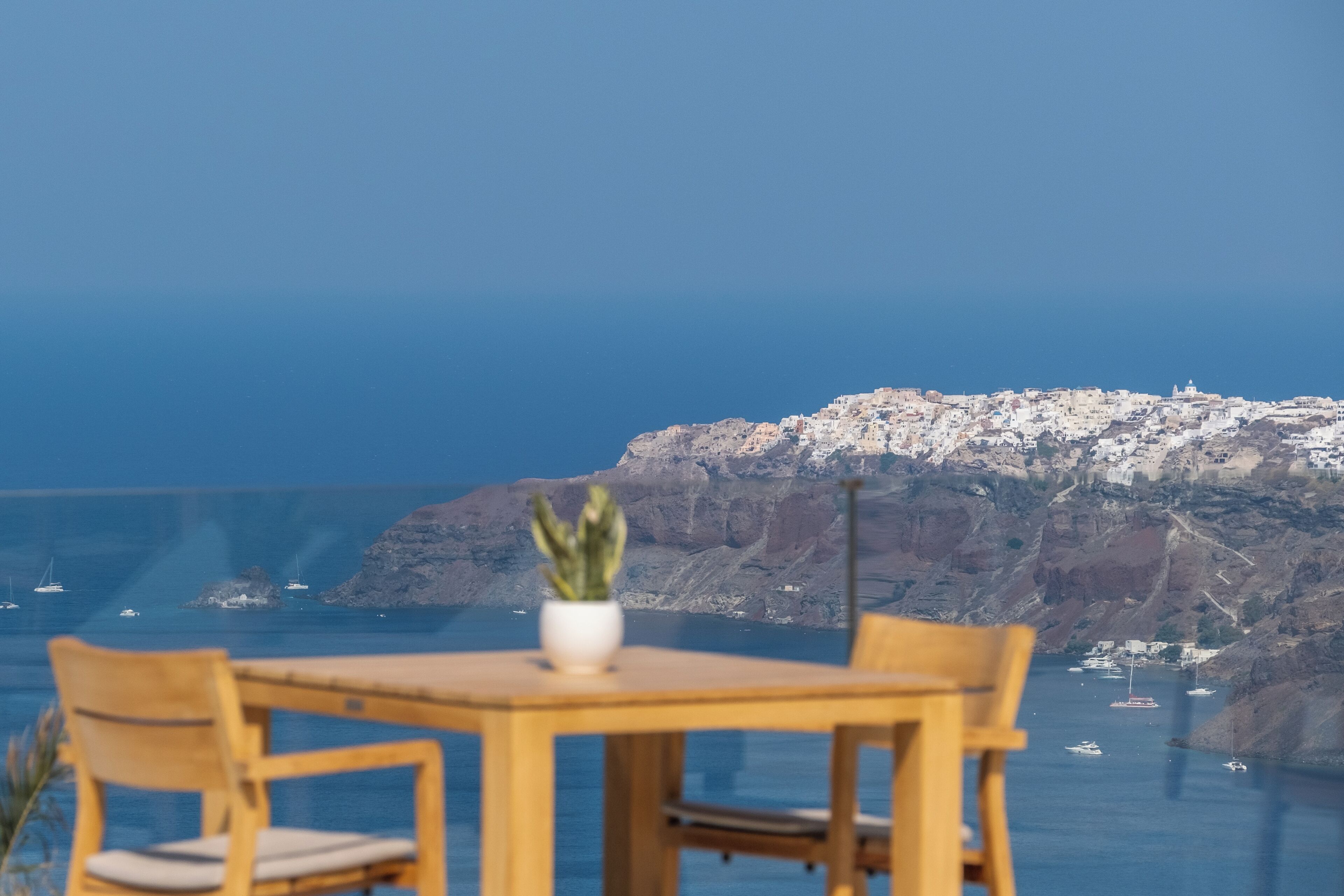 Mesa con sillas y una planta en maceta al frente, vista de un pueblo costero y el mar al fondo.