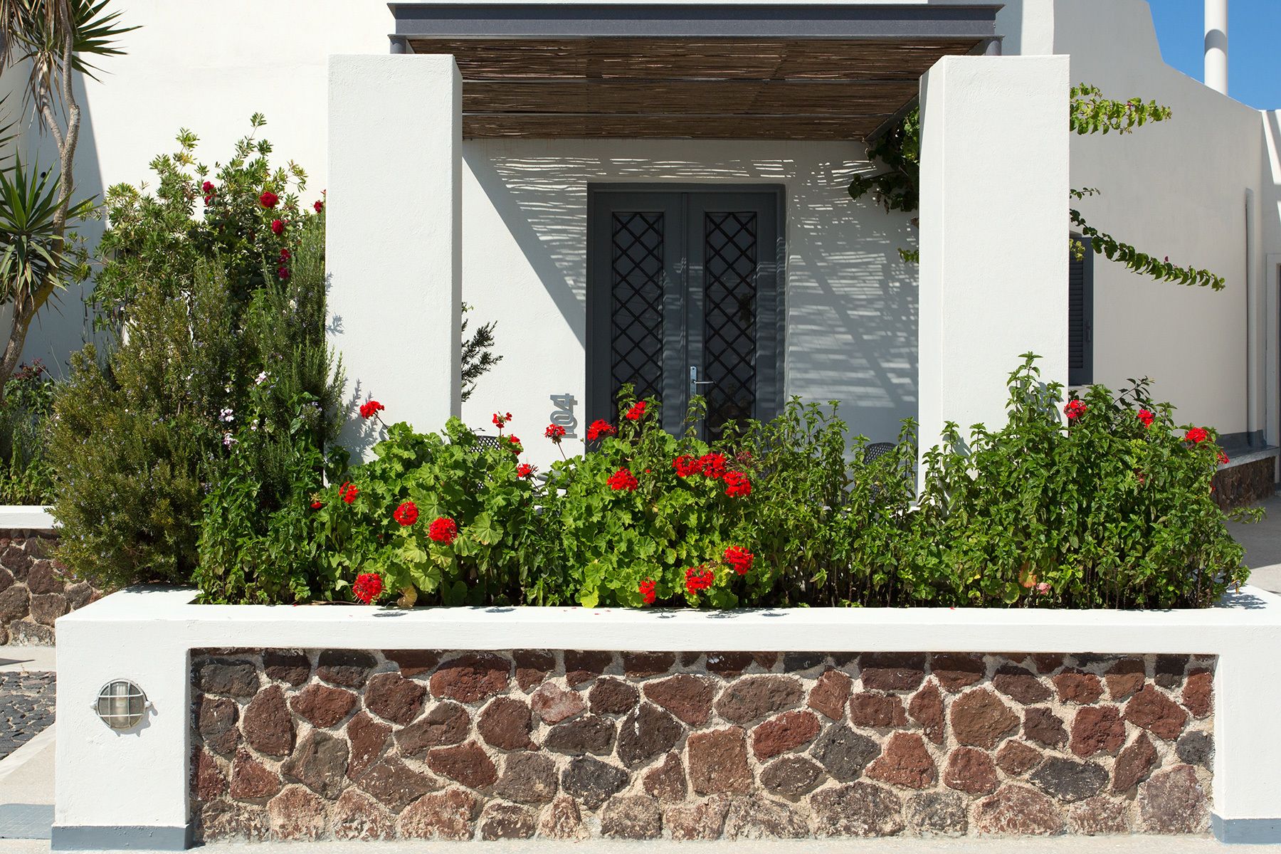 Un edificio moderno con fachada de piedra, una pérgola de madera y flores rojas en primer plano.