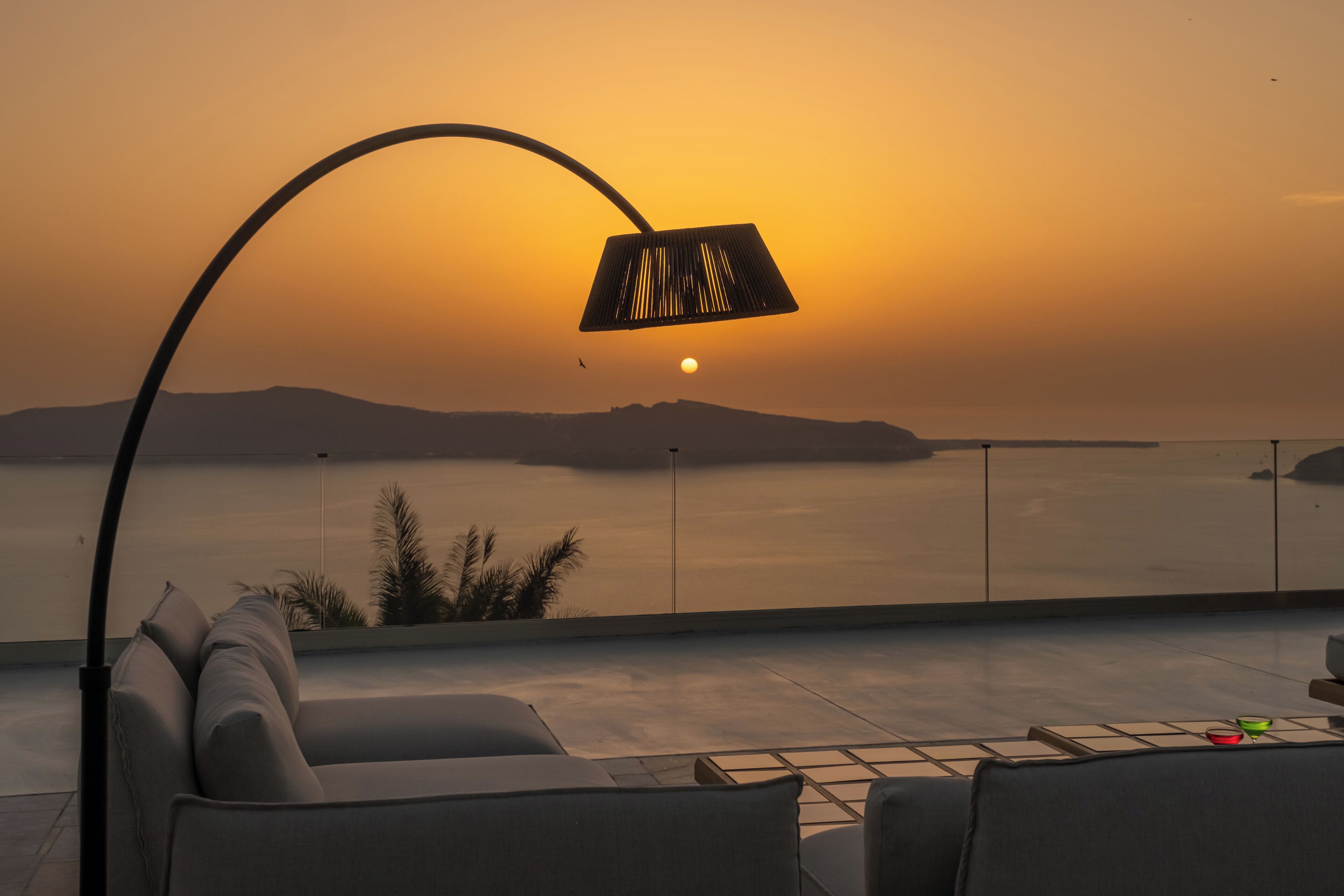A sunset view from a balcony with a lamp, seating, and a railing.