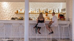 A bar area with white stone walls, a variety of bottles on shelves, and white metal chairs. - Xenones Filotera (Santorini)