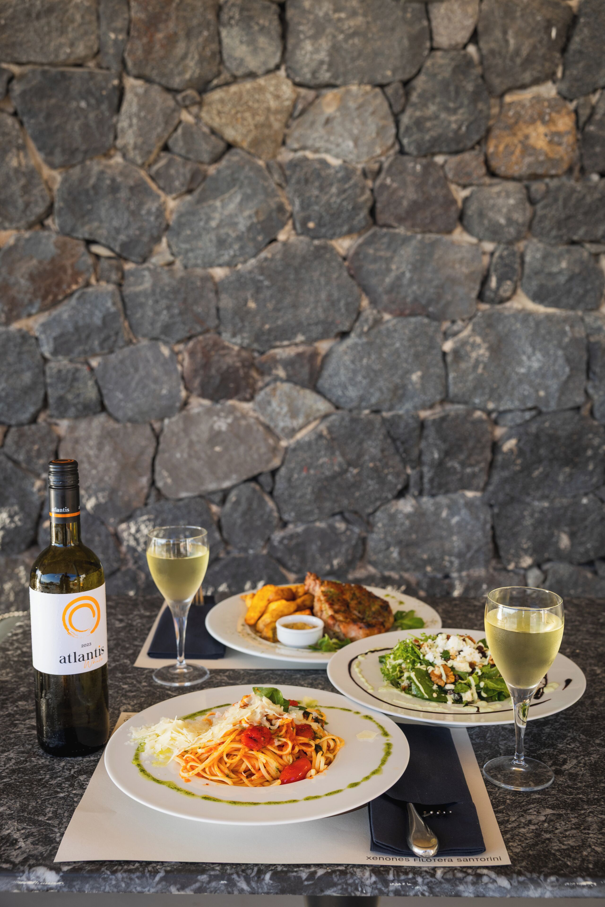 A table with a bottle of wine, two glasses of white wine, and three plates of food.