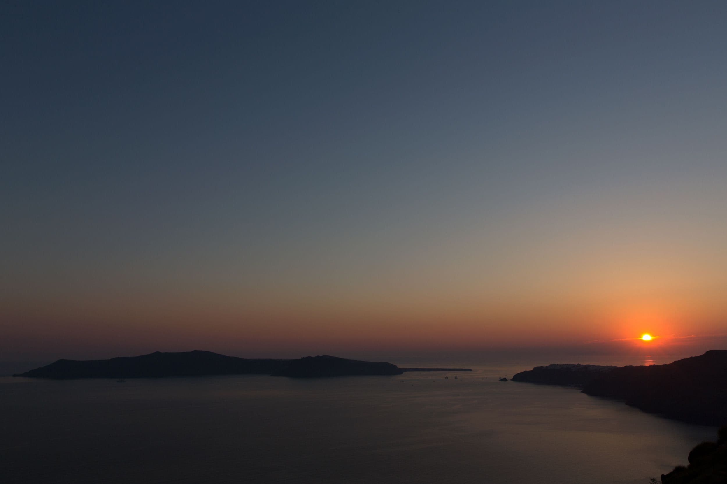 Un coucher de soleil sur la côte, avec des eaux calmes et des reliefs terrestres visibles au loin.
