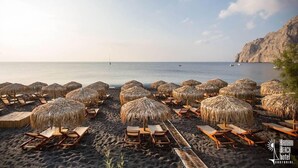 A beach with rows of sun loungers and thatched umbrellas, a calm sea, and a rocky cliff in the background. - Xenones Filotera (Santorini)