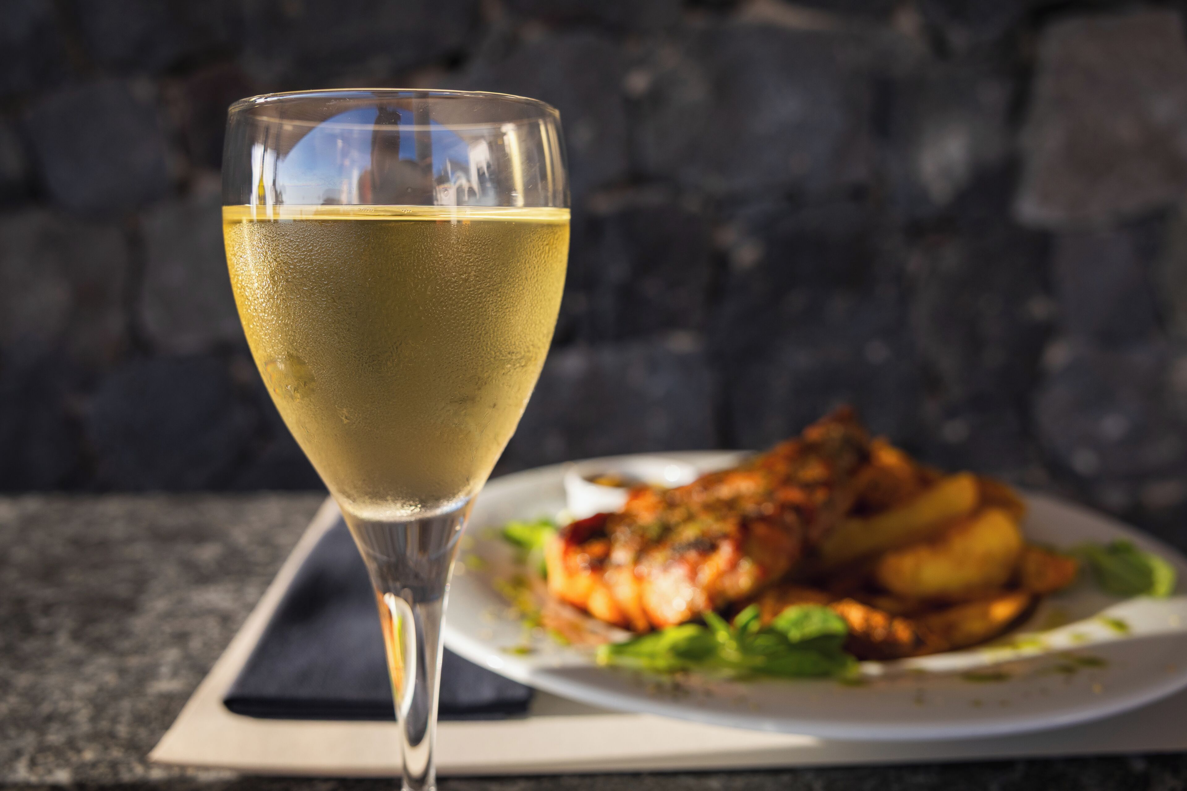 A glass of white wine on a table with a plate of food in the background.