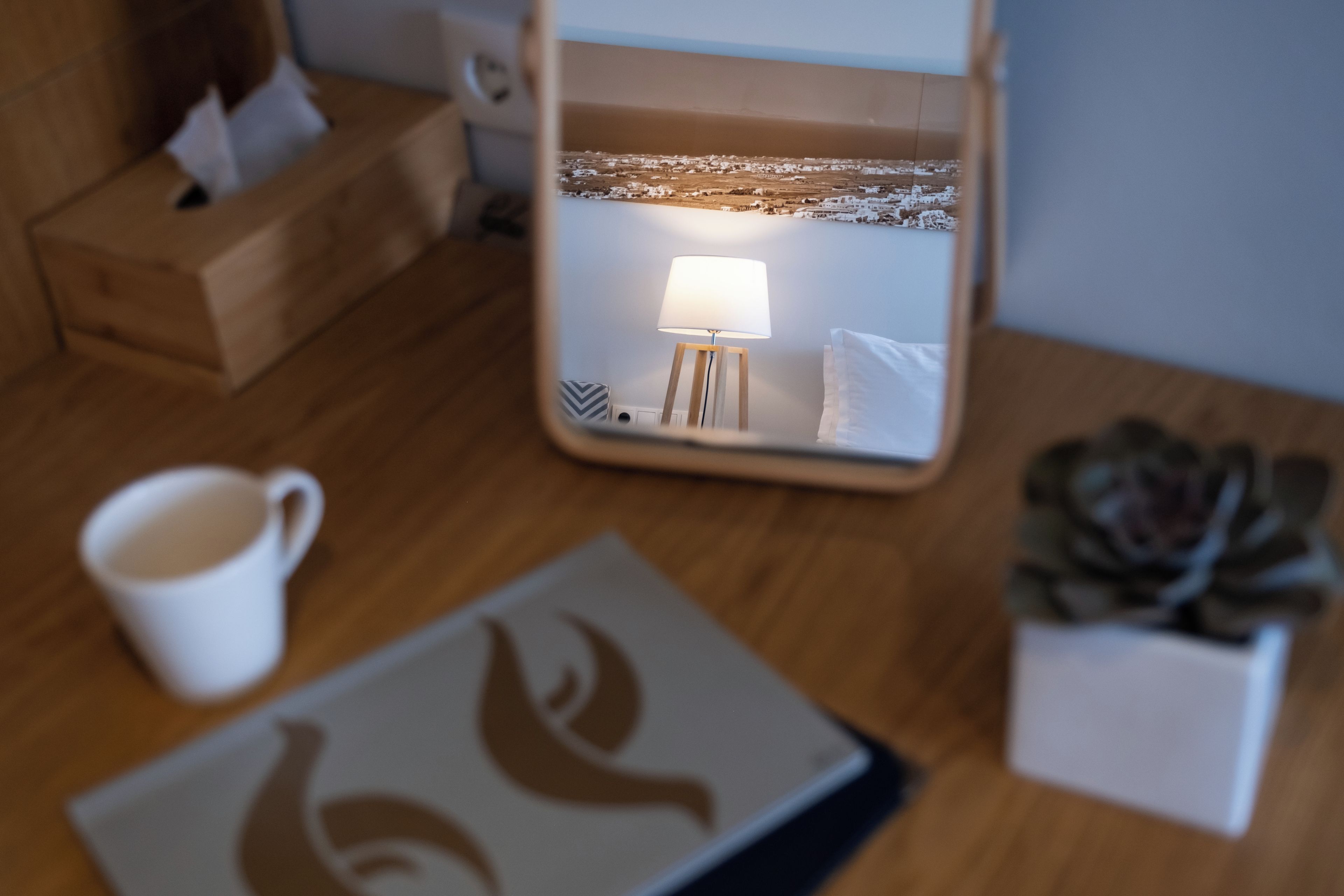 A wooden table with a white mug, a framed picture, a small potted plant, and a mirror reflecting a lamp and a wall.