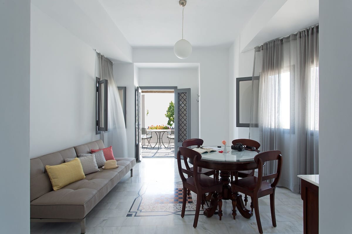 A modern living room with a sofa, dining table, and chairs. There is a view of the outside through an open door.
