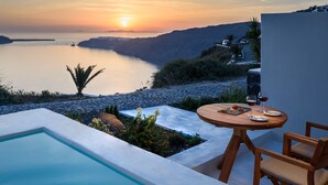 A poolside area with a table and chairs, overlooking a calm body of water and a mountainous landscape during sunset. - Xenones Filotera (Santorini)