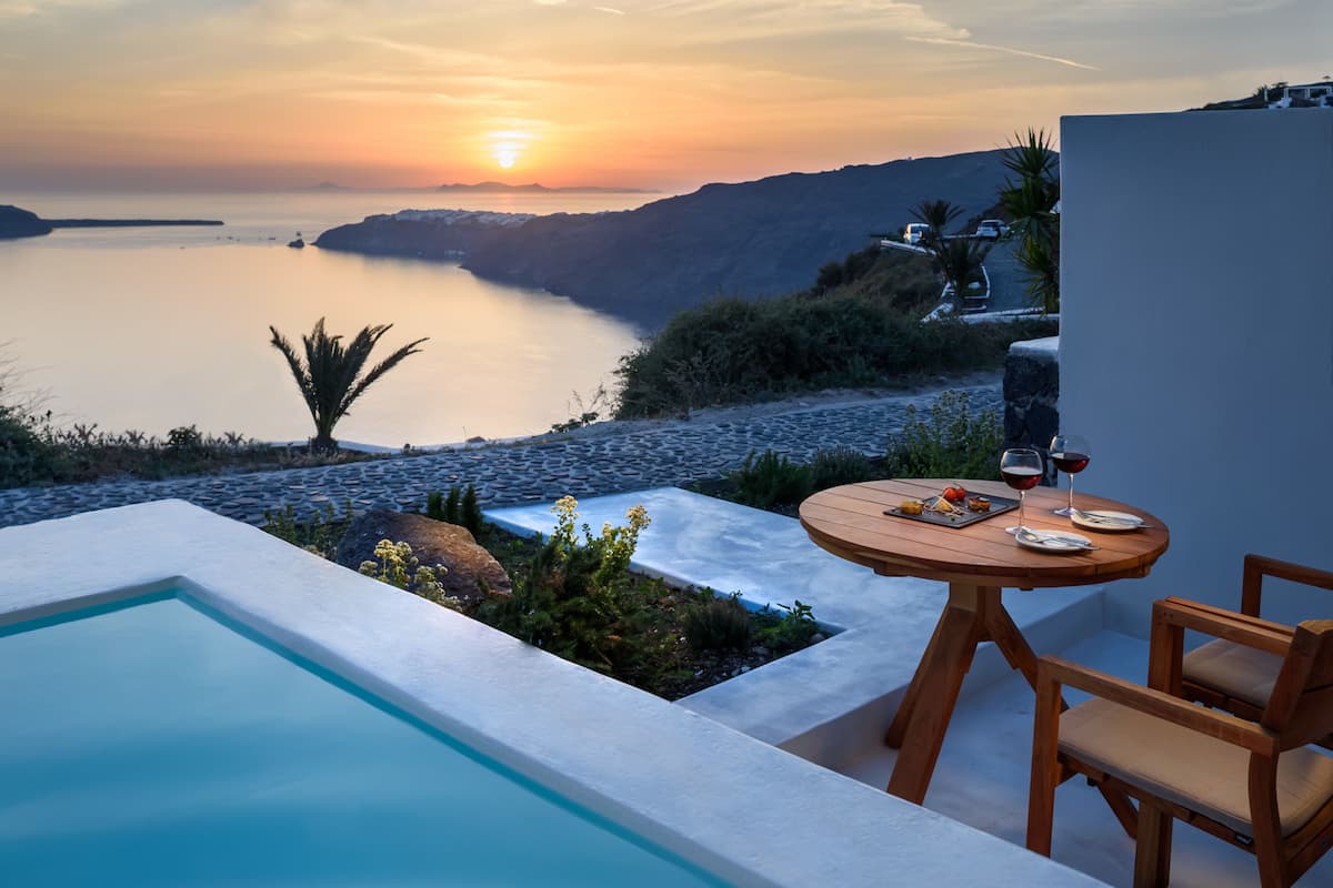 A poolside area with a table and chairs, overlooking a calm body of water and a mountainous landscape during sunset.