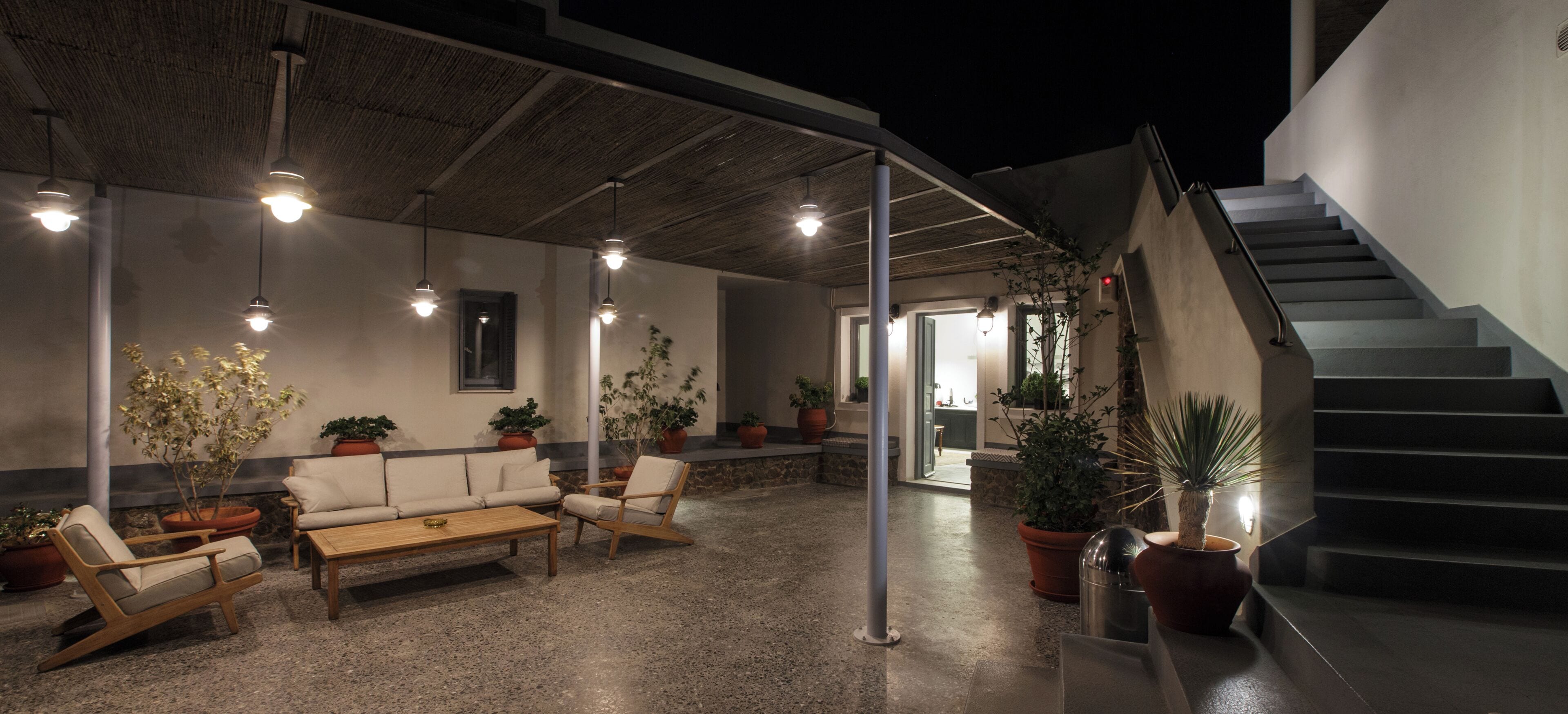 An outdoor seating area with a sofa, chairs, and a table, surrounded by potted plants and under a covered roof.
