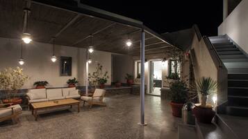 An outdoor seating area with a sofa, chairs, and a table, surrounded by potted plants and under a covered roof.