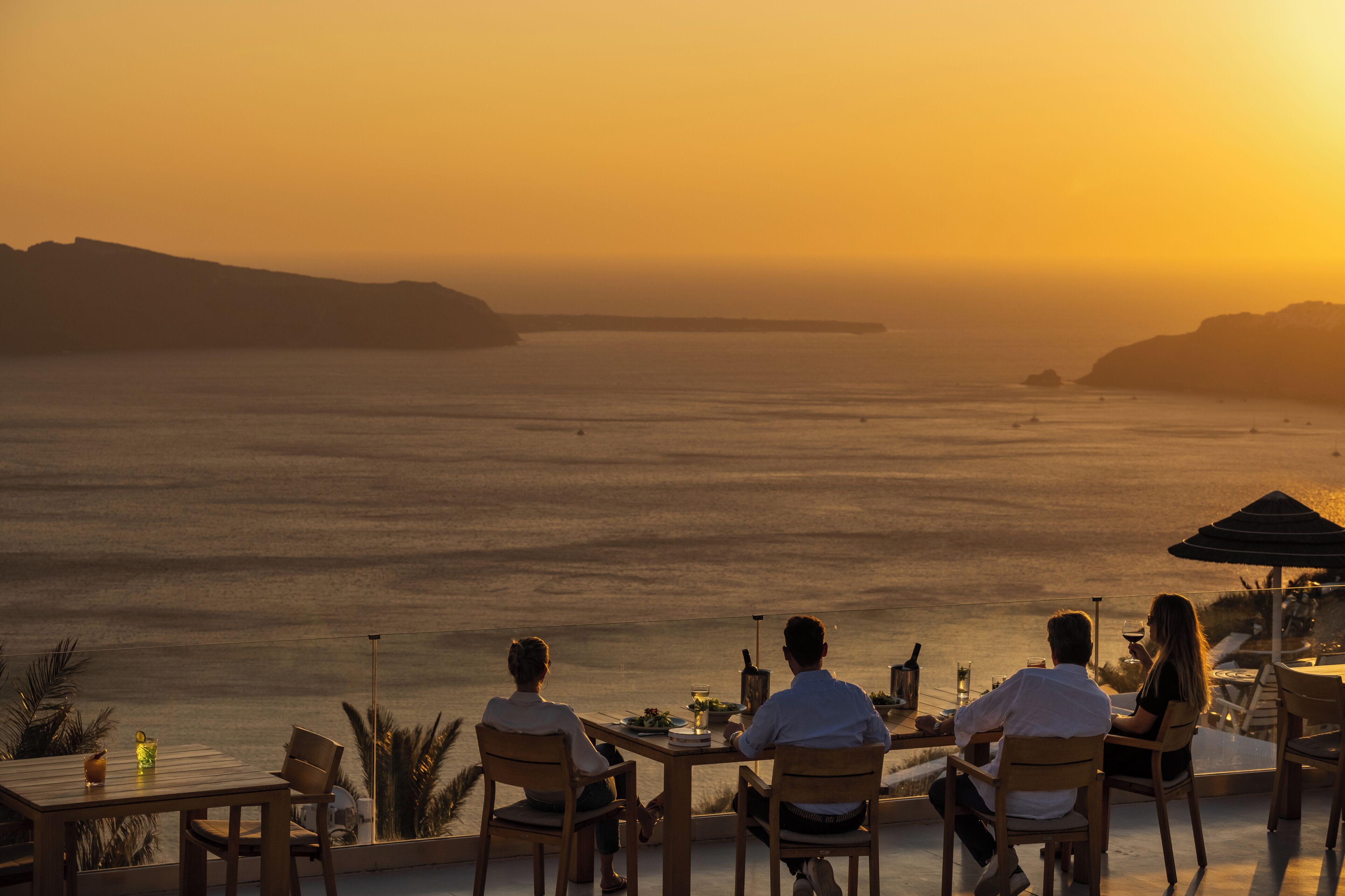 A sunset view from a terrace with a table and chairs, overlooking a body of water and distant landforms.