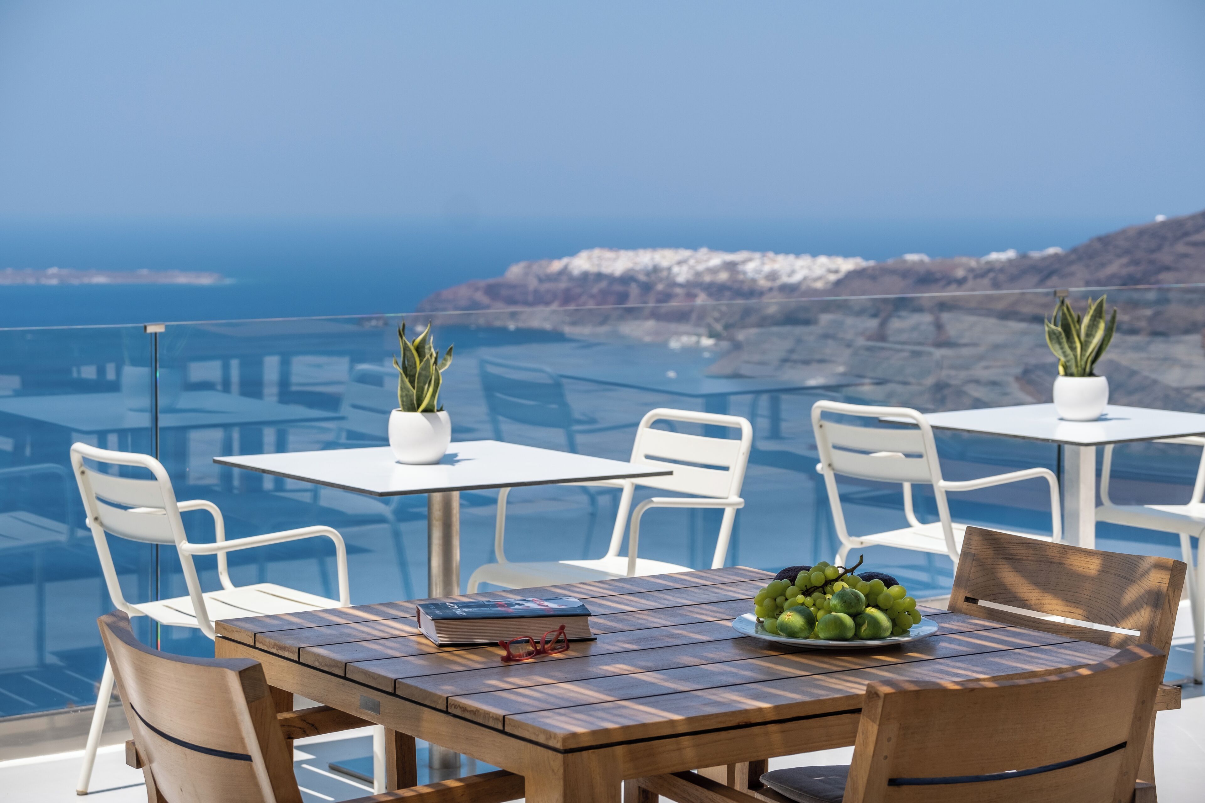 A wooden table with a book and a plate of fruit, white chairs, and potted plants on a balcony overlooking a coastal view.