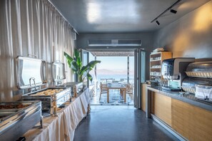A modern kitchen with a buffet setup, a dining area with tables and chairs, and a view of the sea.