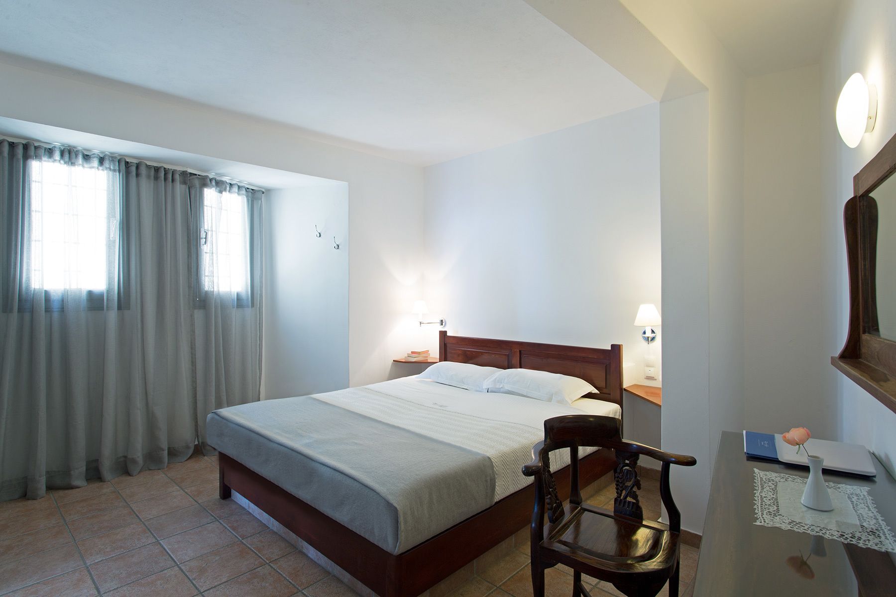 A hotel room with a bed, a chair, a desk with a laptop, and a bedside table with a lamp.