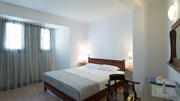 A hotel room with a bed, a chair, a desk with a laptop, and a bedside table with a lamp.
