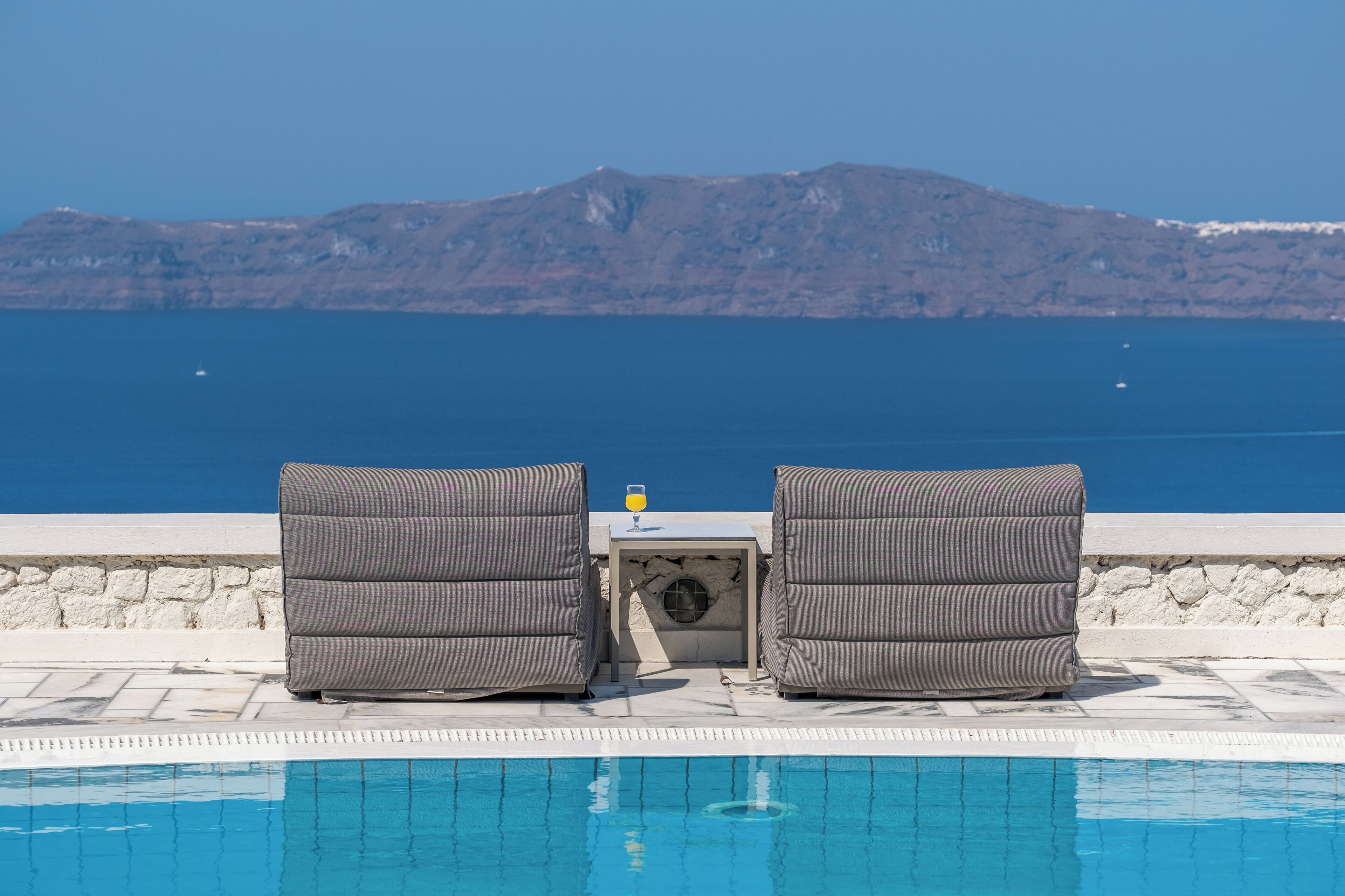 A poolside area with two chairs and a table, overlooking a clear blue sea and a mountainous backdrop.