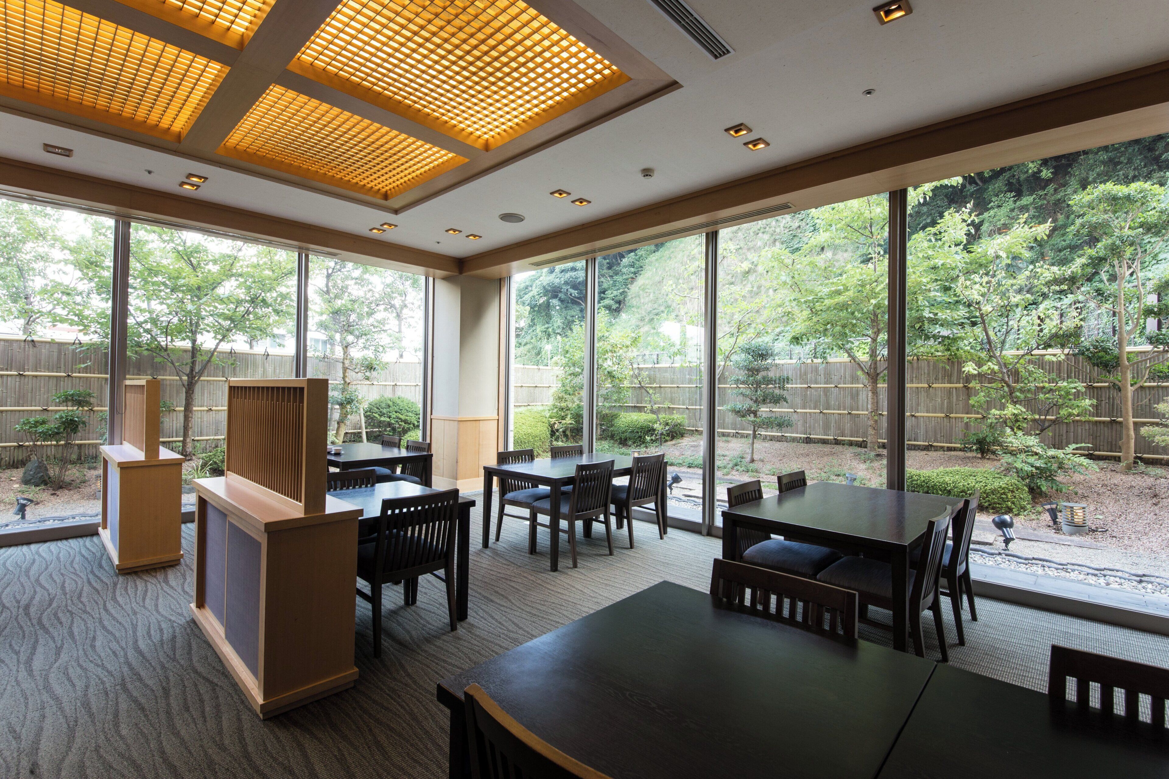 A spacious indoor dining area with large windows, wooden furniture, and a view of a landscaped garden.