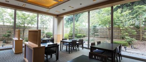 A spacious indoor dining area with large windows, wooden furniture, and a view of a landscaped garden.