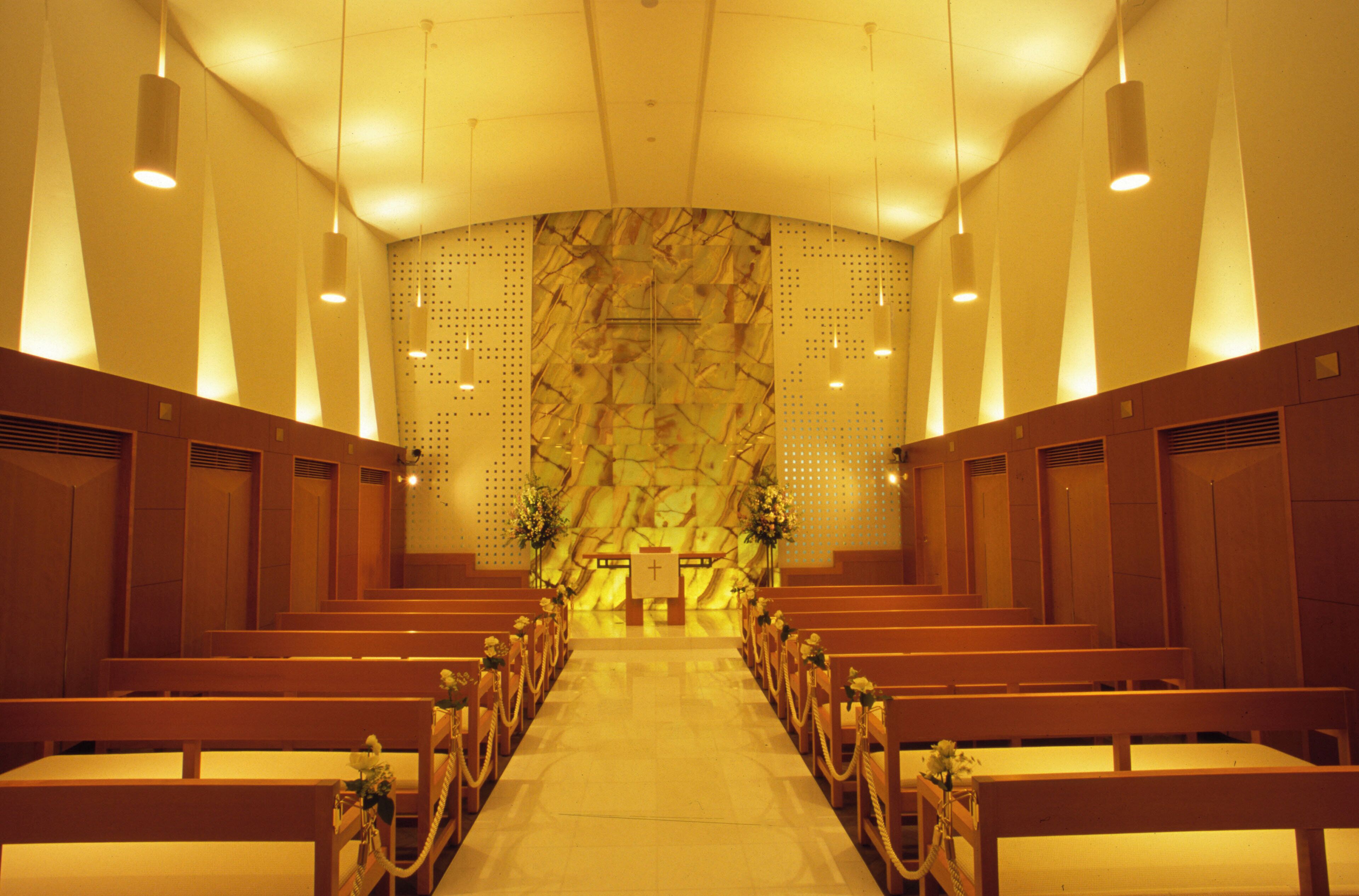 A church interior with pews arranged in rows, a central aisle leading to an altar, and a patterned wall behind the altar.