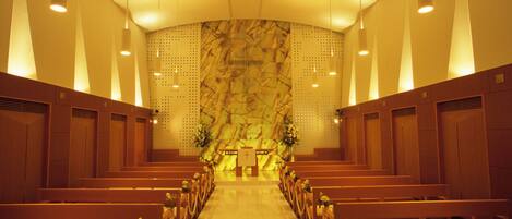 A church interior with pews arranged in rows, a central aisle leading to an altar, and a patterned wall behind the altar.
