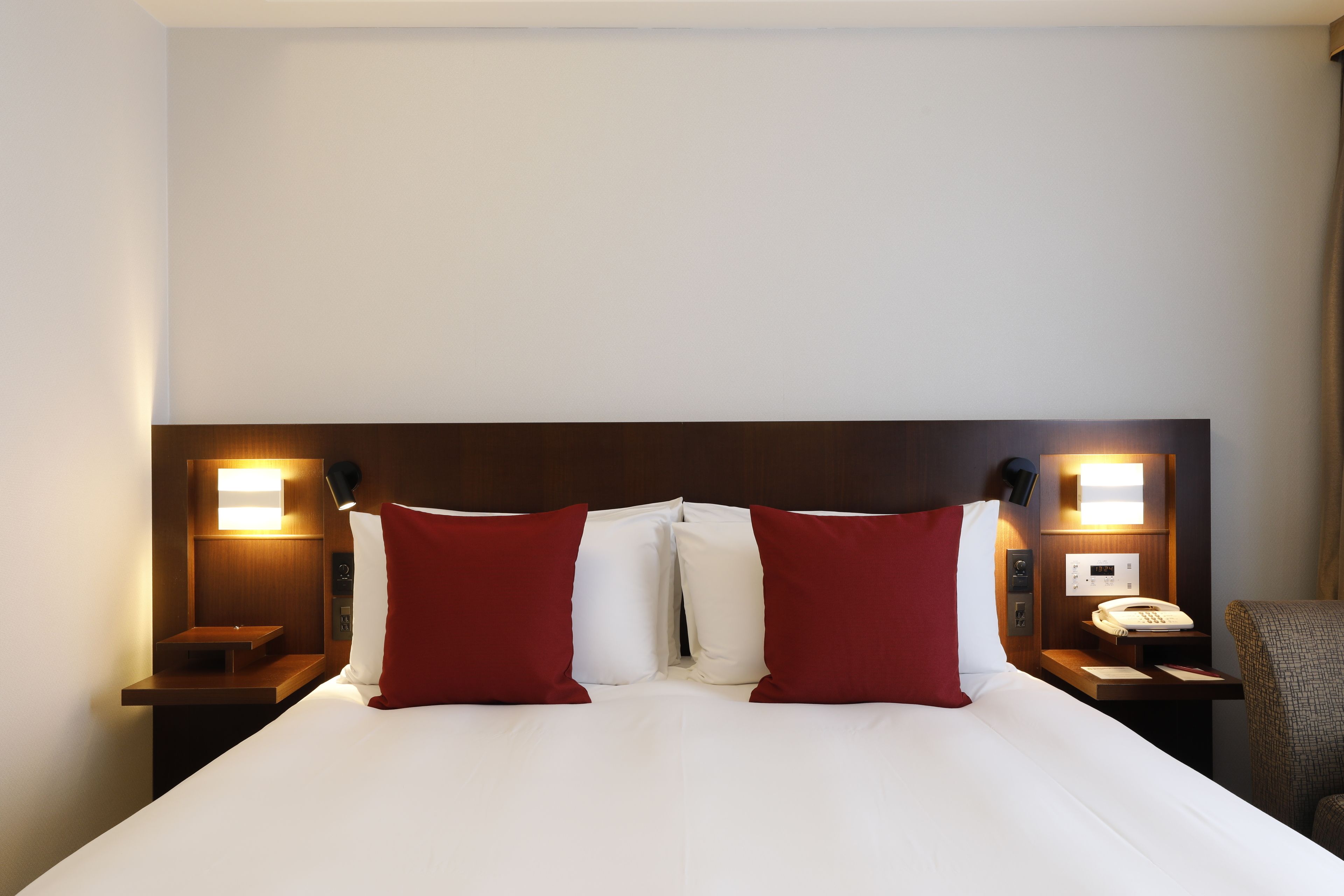 A hotel room with a large bed, two red pillows, a wooden headboard, bedside lamps, a nightstand with a phone, and a chair.