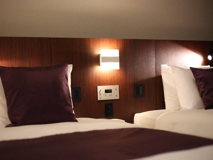 A hotel room with two beds, each with a white mattress and a dark-colored pillow. There is a wall-mounted thermostat between the beds.