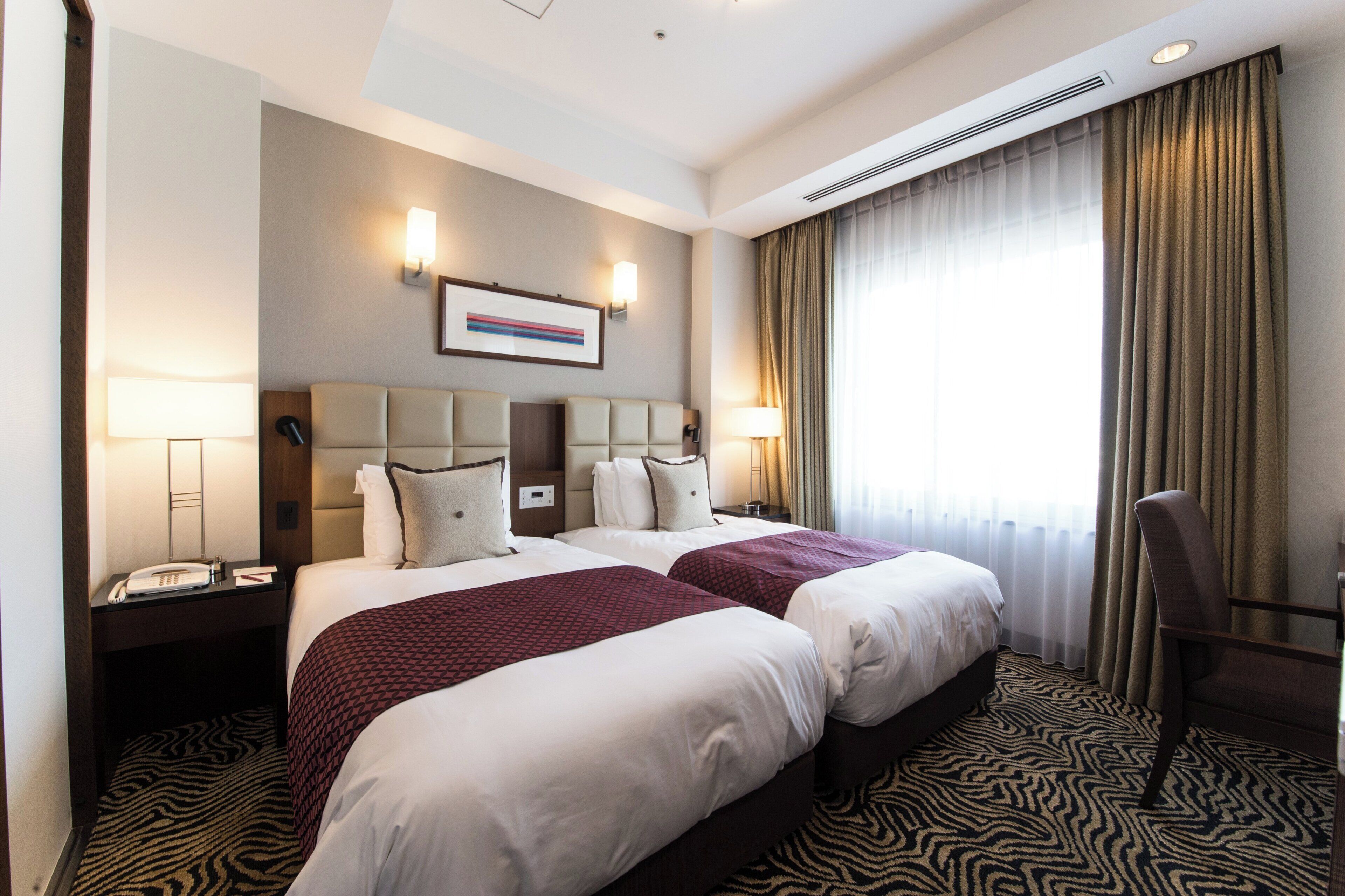 Une chambre d’hôtel avec deux lits, une tête de lit, des lampes de chevet, un téléphone, un tableau encadré et une fenêtre avec des rideaux.