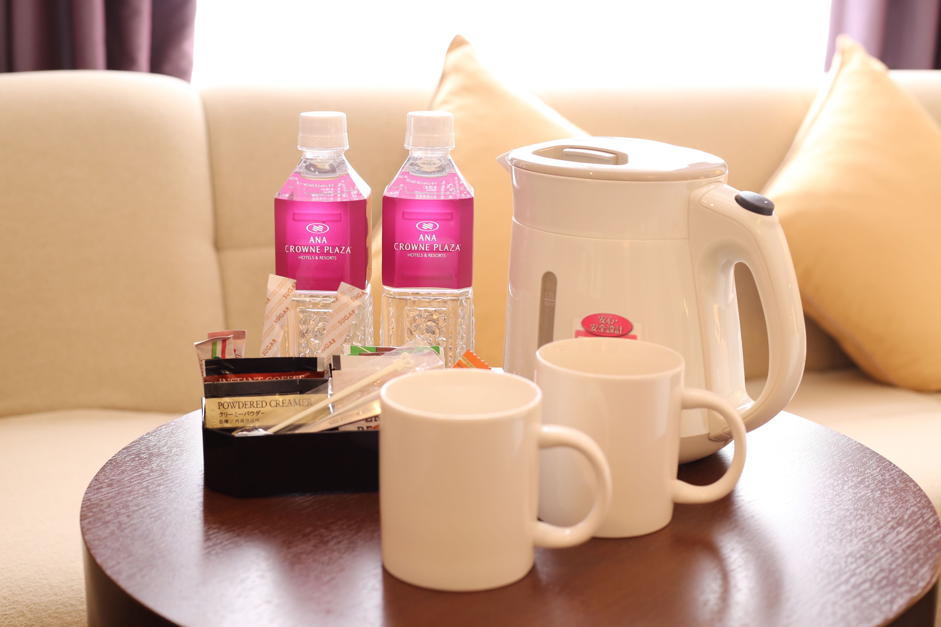 A table with a coffee maker, two bottles of water, a box of creamers, and two white mugs on a round table.