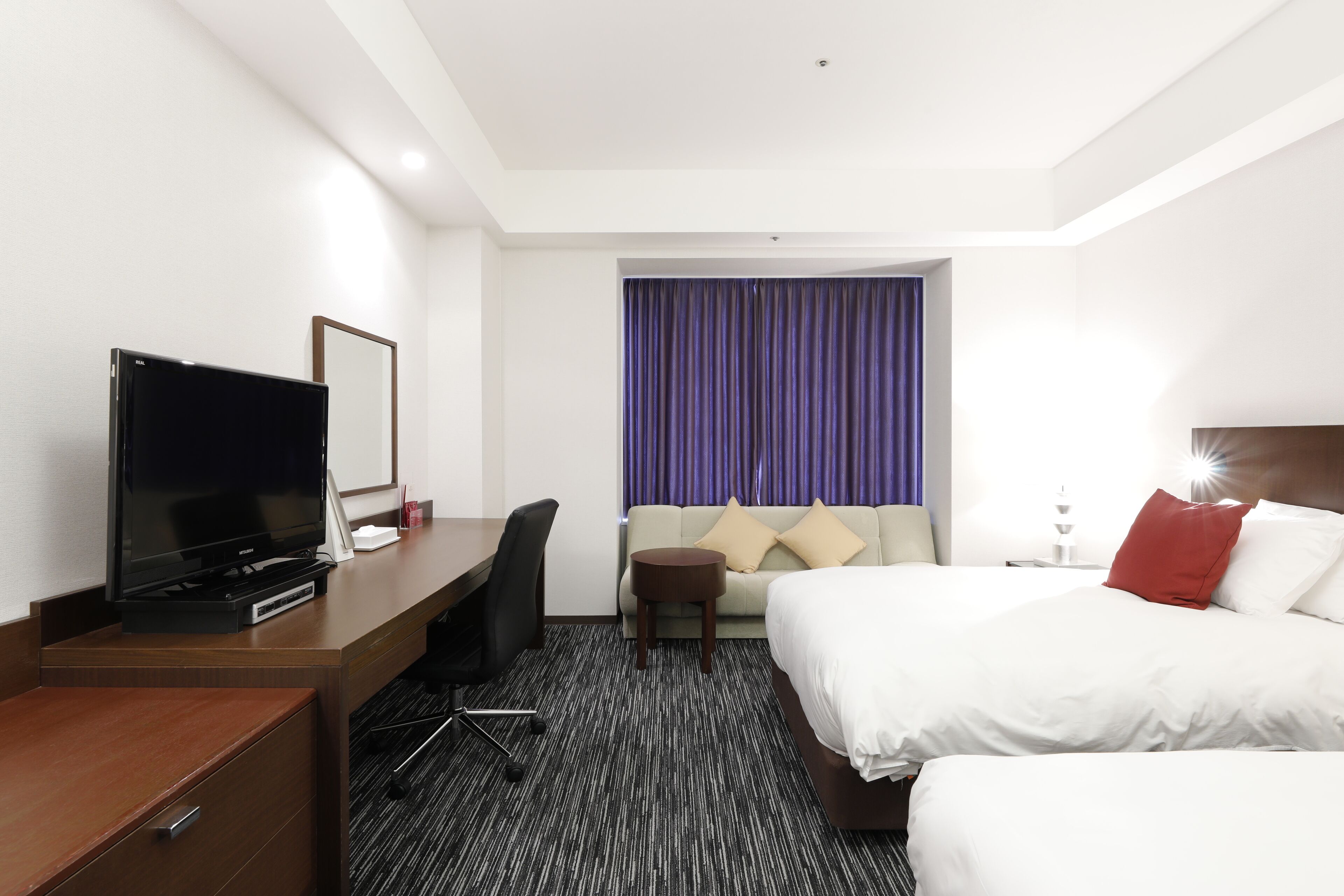 A hotel room with a bed, a desk with a TV, a chair, a small table, and a window with curtains.
