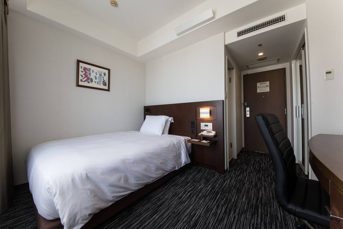 Ein Hotelzimmer mit einem Einzelbett, einem Nachttisch mit Lampe, einem Sessel und einer Tür zu einem anderen Raum.