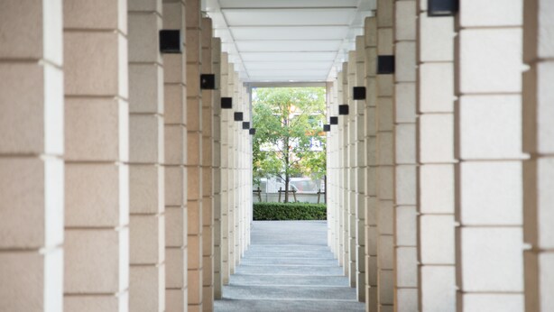 A corridor with a repeating pattern of beige and white tiles, leading to a tree-lined street.