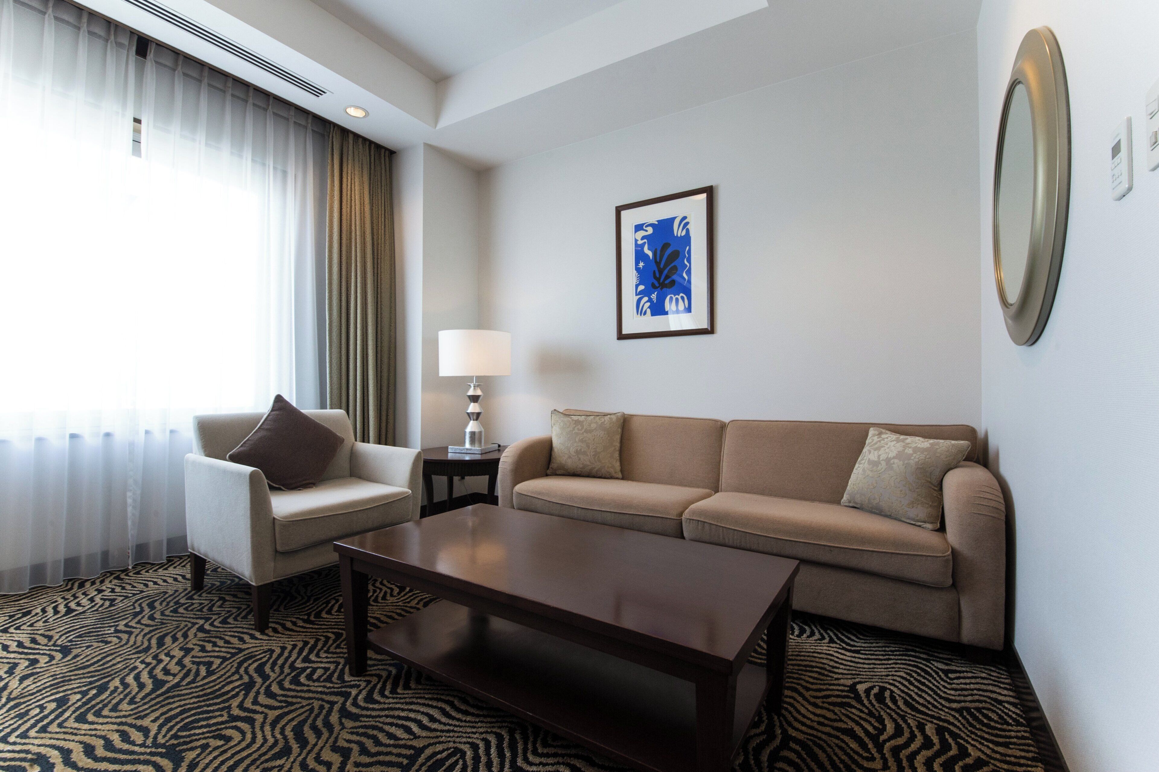 A hotel room with a sofa, armchair, coffee table, and a painting on the wall.