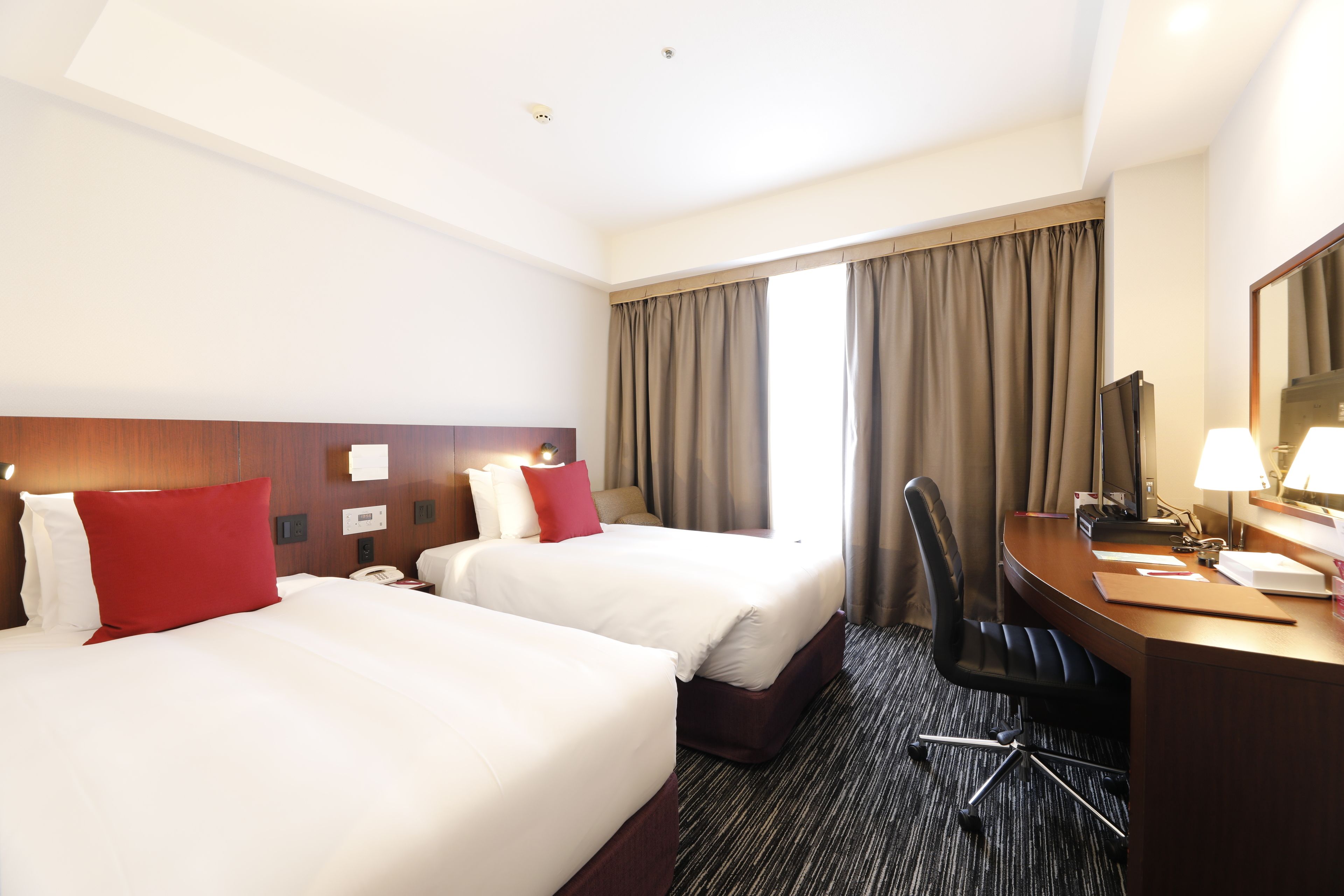 A hotel room with two beds, a desk, a chair, a desk lamp, and a large window with curtains.