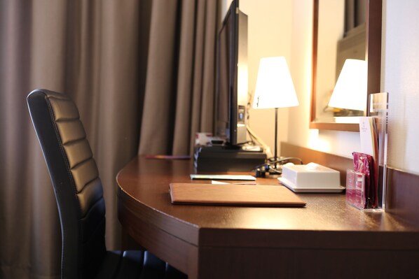 A hotel room with a wooden desk, a chair, a lamp, a mirror, and a bedside table with a tissue box and a glass. - ANA Crowne Plaza Yonago by IHG (Yonago)
