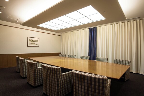 Un salón de conferencias con una larga mesa, seis sillas, un amplio ventanal con persianas y un cuadro en la pared.